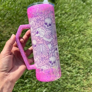 May include: A pink glitter tumbler with a handle, featuring a design of skulls and spiderwebs. The tumbler has a matching pink handle and a black and white patterned straw. The background is blurred green grass.
