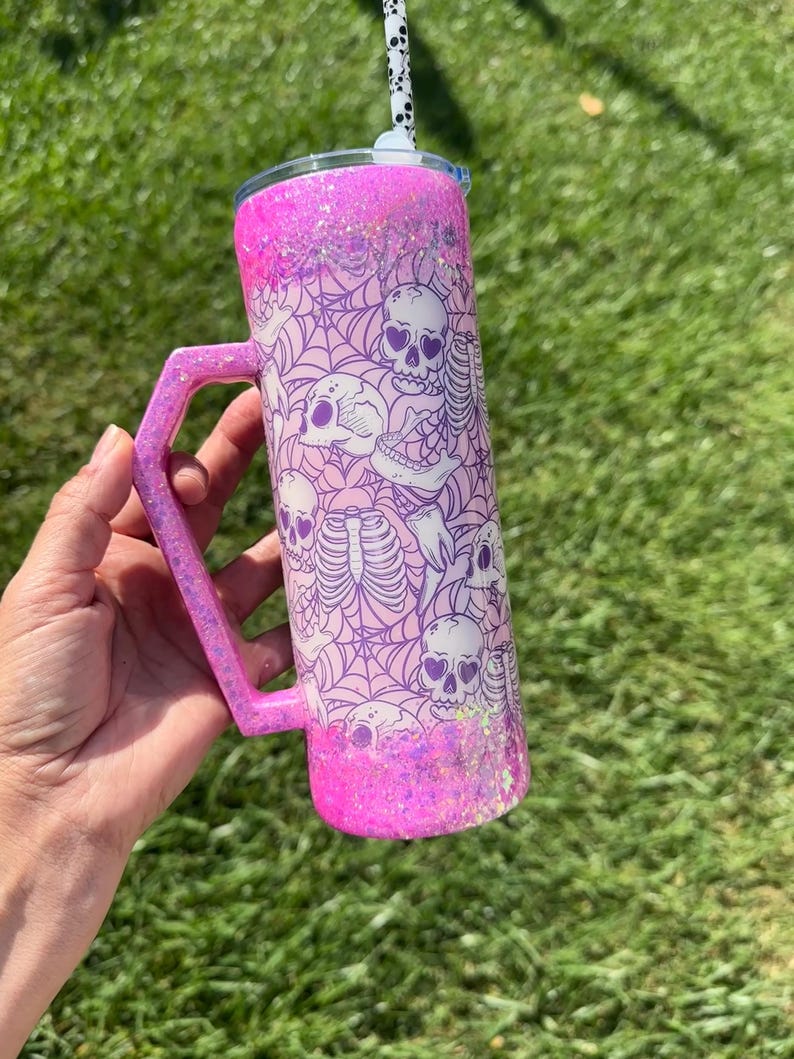 May include: A pink glitter tumbler with a handle, featuring a design of skulls and spiderwebs. The tumbler has a matching pink handle and a black and white patterned straw. The background is blurred green grass.