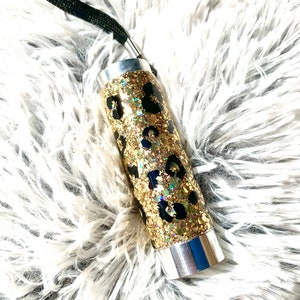 Puede incluir: Un vaso de glitter dorado con un diseño de estampado de leopardo negro. El vaso tiene una tapa plateada y una correa negra.