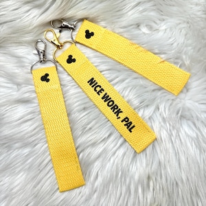 May include: Three yellow fabric wristlet keychains with a textured weave. Each keychain features a small black embroidered mouse head icon and a metal lobster clasp. One keychain has the black embroidered text NICE WORK, PAL on its surface.