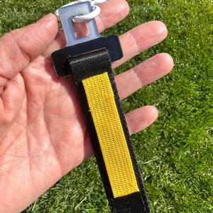 Puede incluir: Un llavero de cintur&oacute;n de seguridad negro y amarillo con un cierre blanco. El llavero tiene una correa negra con una franja central amarilla. El cierre es de pl&aacute;stico blanco y tiene un anillo de metal.