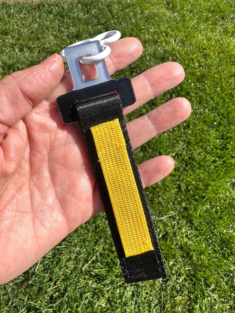 Puede incluir: Un llavero de cintur&oacute;n de seguridad negro y amarillo con un cierre blanco. El llavero tiene una correa negra con una franja central amarilla. El cierre es de pl&aacute;stico blanco y tiene un anillo de metal.