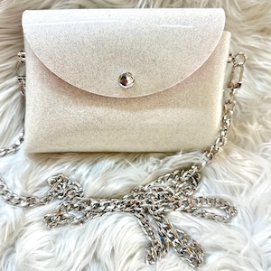 May include: A small white glitter crossbody bag with a silver chain strap. The bag has a flap closure with a silver button.