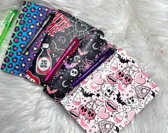 Könnte beinhalten: Eine Sammlung von Reißverschlusstaschen mit verschiedenen Mustern, darunter Leopardenmuster, Spinnennetze und Totenköpfe. Die Taschen sind geöffnet und zeigen Stifte in verschiedenen Farben. Eine Tasche trägt den Text "TEARS OF MY HATERS".