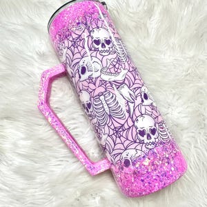 May include: A pink glitter tumbler with a handle, featuring a design of skulls and spiderwebs. The tumbler has a black and white patterned straw and a black lid. The handle and bottom of the tumbler are covered in pink glitter.