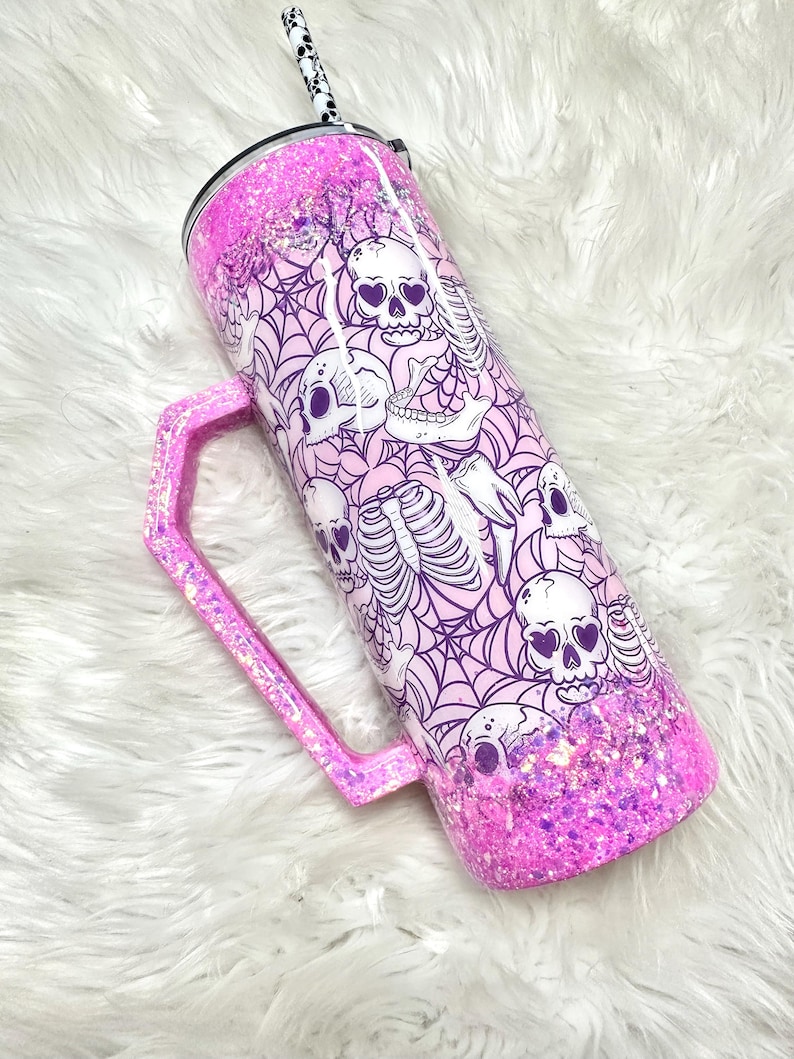 May include: A pink glitter tumbler with a handle, featuring a design of skulls and spiderwebs. The tumbler has a black and white patterned straw and a black lid. The handle and bottom of the tumbler are covered in pink glitter.