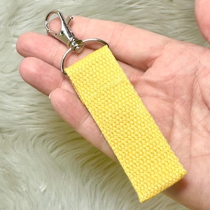 May include: A bright yellow keychain strap with a silver-colored metal clasp and ring. The strap is made of a woven fabric. The keychain is held in a hand.
