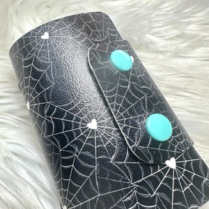 May include: Black leather wallet with a white spiderweb and heart pattern. The wallet has two turquoise buttons.