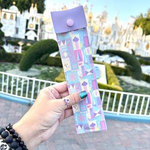 Puede incluir: Una bolsa de tela con estampado morado y azul con una solapa morada y un botón rosa. La bolsa se sostiene en una mano frente a un edificio blanco con un techo de colores.