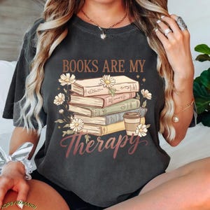 May include: Dark gray t-shirt with the text "BOOKS ARE MY Therapy" in brown and burgundy script. The graphic features a stack of books, daisies, and a coffee cup. The shirt is a comfortable, casual style.