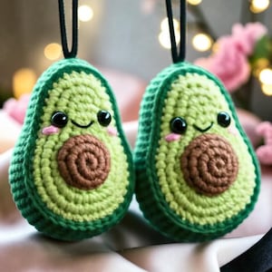 Könnte beinhalten: Zwei handgefertigte Häkel-Avocado-Anhänger mit hellgrüner Mitte, dunkelgrünem Rand und braunem Kern. Jede Avocado hat schwarze Sicherheitsaugen, ein aufgesticktes Lächeln und rosa Wangen. An der Oberseite ist jeweils eine schwarze Kordel befestigt.