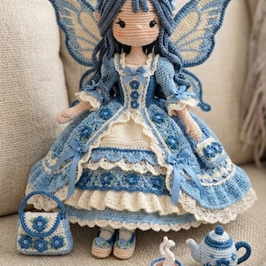 Könnte beinhalten: Eine handgefertigte Häkel-Feenpuppe mit langem blauem Haar und großen Flügeln, die ein mehrlagiges blau-cremefarbenes Kleid mit Blumenakzenten trägt. Die Puppe wird von einer passenden kleinen Häkeltasche, einer Teetasse und einer Teekanne begleitet.