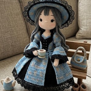 Könnte beinhalten: Eine gehäkelte Puppe mit grauem Haar trägt ein blau-schwarzes Kleid mit einem passenden breitkrempigen Hut, der mit schwarzer Spitze verziert ist. Die Puppe hält eine kleine Teetasse, begleitet von einer Miniatur-Teekanne, einem Teller mit Keksen, einer Handtasche und braunen Stiefeln.