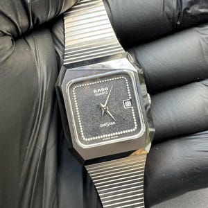 Puede incluir: Un reloj Rado Diastar Quartz vintage con una esfera cuadrada negra y agujas plateadas. El reloj tiene una pantalla de fecha a las 3 en punto y una correa de metal plateado. La esfera del reloj está rodeada por un borde de cuentas plateadas.