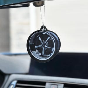 Puede incluir: Un ambientador de coche negro con un diseño de rueda. El ambientador redondo tiene un gráfico de rueda gris y plateado. Cuelga de un hilo sujeto al espejo retrovisor de un coche.