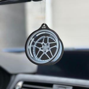 May include: A black and gray car air freshener with a wheel design. The air freshener is round with a detailed illustration of a car wheel in shades of gray and silver. The air freshener is hanging from a string.