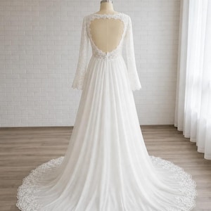 May include: A white floor-length gown featuring long lace sleeves, a heart-shaped open back design, and a flowing skirt with a lace-trimmed hemline. The bodice is detailed with intricate lace patterns that extend down the long sleeves.