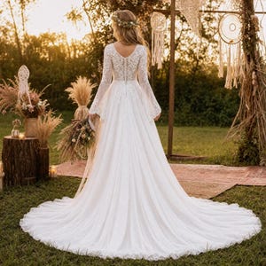 May include: A floor-length white lace wedding dress featuring long bell sleeves, a V-neckline, a fitted bodice with a button-down back, and a flowing skirt with a train. The dress is adorned with intricate floral lace patterns throughout the bodice.