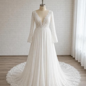 May include: A floor-length white wedding dress featuring a deep V-neckline, long lace sleeves, and a fitted bodice. The skirt is made of flowing fabric with a wide lace-trimmed hem that extends into a circular train, displayed on a mannequin.