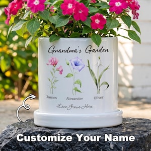 May include: White ceramic planter with the text "Grandma's Garden" and floral illustrations. The planter has a matching saucer and is filled with vibrant pink flowers. The text "Customize Your Name" is at the bottom.