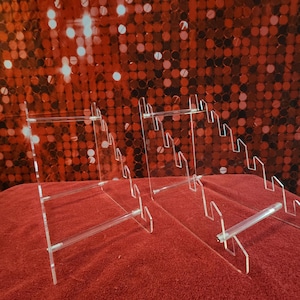 May include: Clear acrylic display stands with multiple tiers and horizontal bars. The stands are on a red surface with a sparkly red background. The stands are designed to hold and showcase items.
