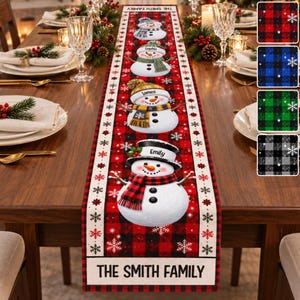 May include: A long rectangular table runner featuring a red and black buffalo plaid pattern with white snowflake accents. The runner displays four illustrated snowmen with personalized names and a bottom border reading THE SMITH FAMILY.