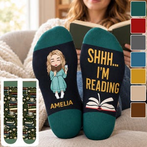 May include: A pair of dark-coloured socks with teal cuffs. One sock features a cartoon illustration of a person reading a book with the name AMELIA printed below. The other sock displays the text SHHH... I'M READING above a graphic of an open book.