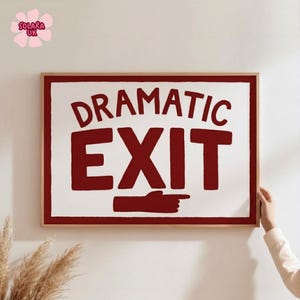 May include: A rectangular wall sign in a light wood frame featuring a white background with a dark red border. The text DRAMATIC EXIT is printed in bold dark red capital letters, with a dark red hand icon pointing to the right below the text.