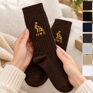 May include: A pair of dark brown ribbed crew socks featuring an embroidered yellow graphic of two figures and the text H heart S. The socks have a vertical ribbed texture and are displayed alongside a color palette of eight additional solid color options.