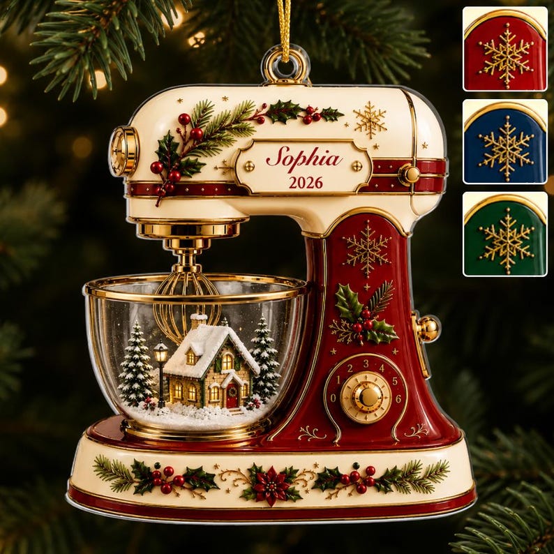 May include: A cream and deep red stand mixer ornament featuring gold accents, holly, and berry motifs. The clear glass bowl contains a miniature snow-covered house and pine trees. A plaque displays the name Sophia and the year 2026.