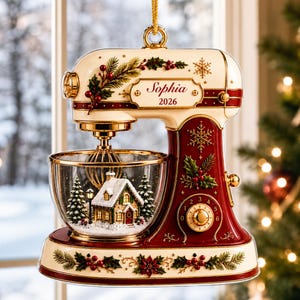 May include: A cream and deep red stand mixer Christmas ornament featuring gold accents, holly, and snowflake patterns. The mixing bowl contains a miniature snow-covered house and pine trees. A plaque on the side displays the text Sophia 2026.