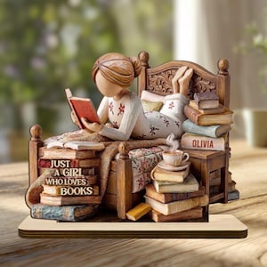Peut inclure: Une figurine représentant une personne allongée sur un lit en train de lire, vêtue d'un pyjama blanc à motifs floraux rouges. Des piles de livres entourent le lit, avec le texte "JUST A GIRL WHO LOVES BOOKS" et "OLIVIA" sur des livres.