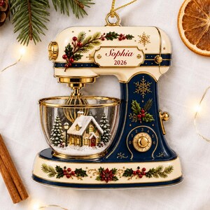 May include: A cream and navy blue stand mixer ornament featuring gold accents, holly, and berry motifs. The mixing bowl contains a miniature snow-covered house and pine trees. A plaque on the side displays the name Sophia and the year 2026.