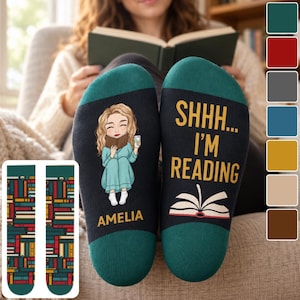 May include: A pair of black and dark green socks featuring a cartoon illustration of a person reading a book, the text "SHHH... I'M READING" above an open book graphic, and the name "AMELIA" printed below the figure, with a book pattern on the cuffs.