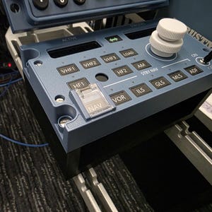 Pode incluir: Um painel de controle de rádio para simulador de voo azul com display digital, botão giratório branco, interruptor preto e vários botões rotulados como VHF1, VHF2, VHF3, LOAD, AM, HF2, HF1, VOR, ILS, GLS, MLS, ADF e um botão NAV.