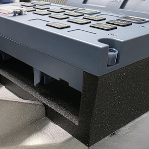 Puede incluir: Primer plano de un panel de control gris con múltiples botones negros y un componente de plástico transparente. El panel está sobre una base negra texturizada y una estructura de soporte plateada.