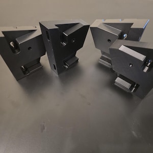 Op de afbeelding: Vier zwarte, geometrische metalen objecten zijn gerangschikt op een donker oppervlak. Elk object heeft een driehoekige vorm met schuine randen en diverse uitsparingen. De objecten lijken precisiebewerkte onderdelen te zijn.