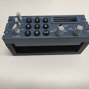 May include: A gray and black aircraft control panel with multiple buttons, knobs, and a small display screen. The panel has labels such as "ATC," "FAIL," and numbers 1 through 7, along with other text. The panel is designed with a rectangular shape and a black base.