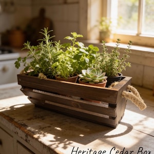 May include: A rectangular dark brown wooden crate with thick jute rope handles on each side. Inside the crate are several small potted plants, including herbs and a succulent. The text "Heritage Cedar Box" is printed in black on the surface.