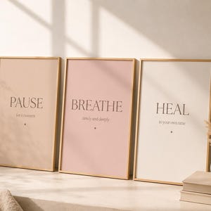 May include: Three rectangular framed wall art prints with light wood frames. The first print is beige with the text PAUSE for a moment. The second is dusty pink with the text BREATHE slowly and deeply. The third is cream with the text HEAL in your own time.