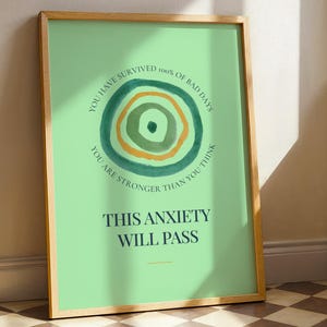 May include: A framed art print with a light green background and a wooden frame. The print features a watercolor design of concentric circles in green and yellow. Text reads: "YOU HAVE SURVIVED 100% OF BAD DAYS. YOU ARE STRONGER THAN YOU THINK. THIS ANXIETY WILL PASS."