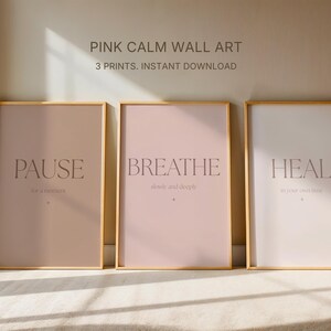 Op de afbeelding: Drie ingelijste wandkunstprints met gouden frames. De prints hebben een roze en witte achtergrond met de woorden "PAUSE", "BREATHE" en "HEAL" in een schreefletter. De tekst "PINK CALM WALL ART" en "3 PRINTS. INSTANT DOWNLOAD" staat bovenaan.