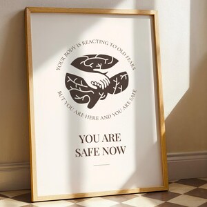 May include: Framed art print with a light background and a wooden frame. The print features a brown illustration of a hand holding a brain, with text that reads "YOUR BODY IS REACTING TO OLD FEARS BUT YOU ARE HERE AND YOU ARE SAFE YOU ARE SAFE NOW."