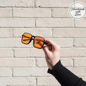 Puede incluir: Gafas de sol con montura negra y lentes de color naranja, sostenidas contra una pared de ladrillos blancos. Las monturas cuadradas y las lentes son el punto focal, con el texto "Handmade with Love" en un c&iacute;rculo en el fondo.