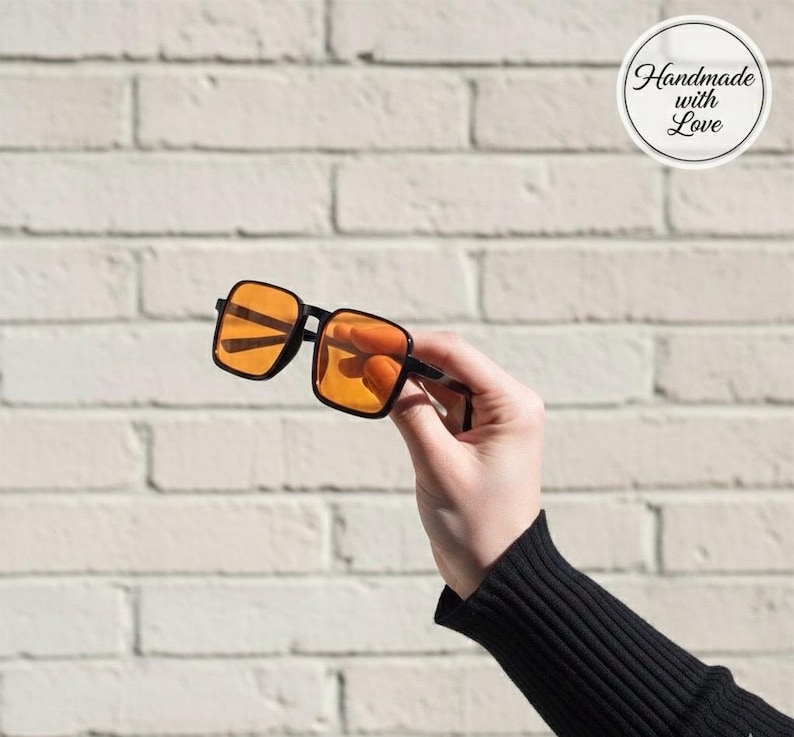 Puede incluir: Gafas de sol con montura negra y lentes de color naranja, sostenidas contra una pared de ladrillos blancos. Las monturas cuadradas y las lentes son el punto focal, con el texto "Handmade with Love" en un c&iacute;rculo en el fondo.