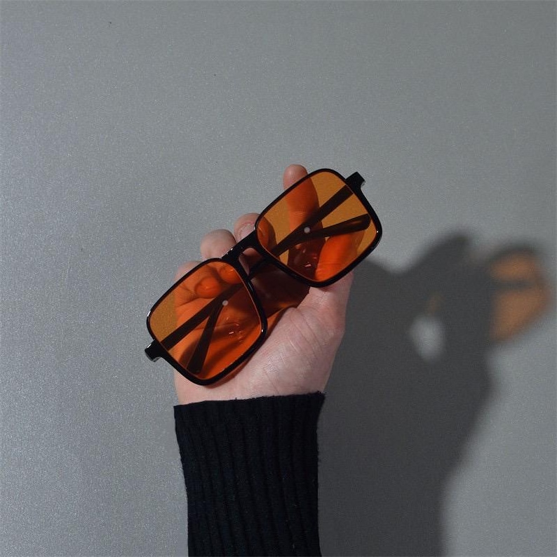Puede incluir: Gafas de sol con montura negra y lentes naranjas. Las monturas cuadradas se sostienen en una mano sobre un fondo gris. Las lentes son de color naranja brillante y las patillas de las gafas son finas y negras.