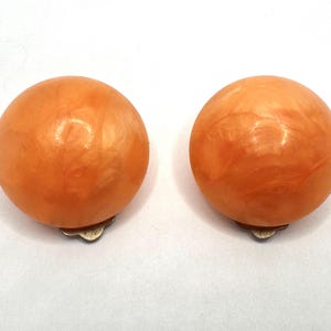 May include: Pair of orange clip-on earrings. Each earring is a round, marbled sphere with a silver-colored clip. The earrings have a warm, peachy hue and a glossy finish, suitable for adding a pop of color to any outfit.