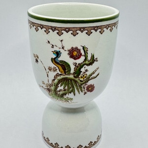 May include: White ceramic egg cup with a decorative design featuring a colorful bird perched on a branch with flowers. The rim is trimmed with green and brown decorative patterns. The egg cup has a pedestal base.