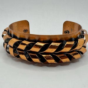 May include: A copper cuff bracelet with a woven design. The bracelet features a wide band with a hammered texture and a series of interwoven copper ribbons. The metal has a warm, reddish-brown hue and a polished finish. The bracelet is open at the back.