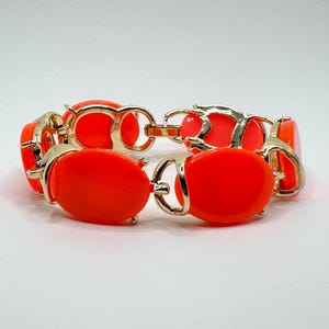 May include: A vibrant orange bracelet with gold-tone metal links. The bracelet features oval-shaped orange resin stones set within the gold links. A bold and eye-catching piece of jewelry.